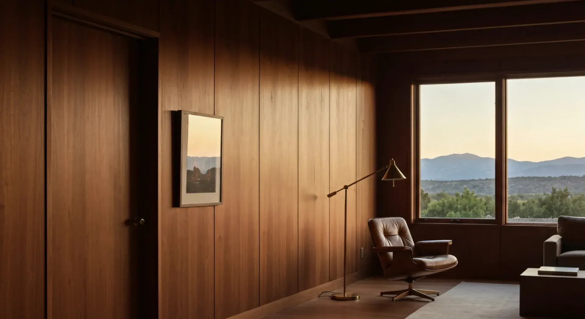 A leather lounge chair and brass floor lamp beside a window at dusk in a walnut-paneled room, hills fading in the distance.