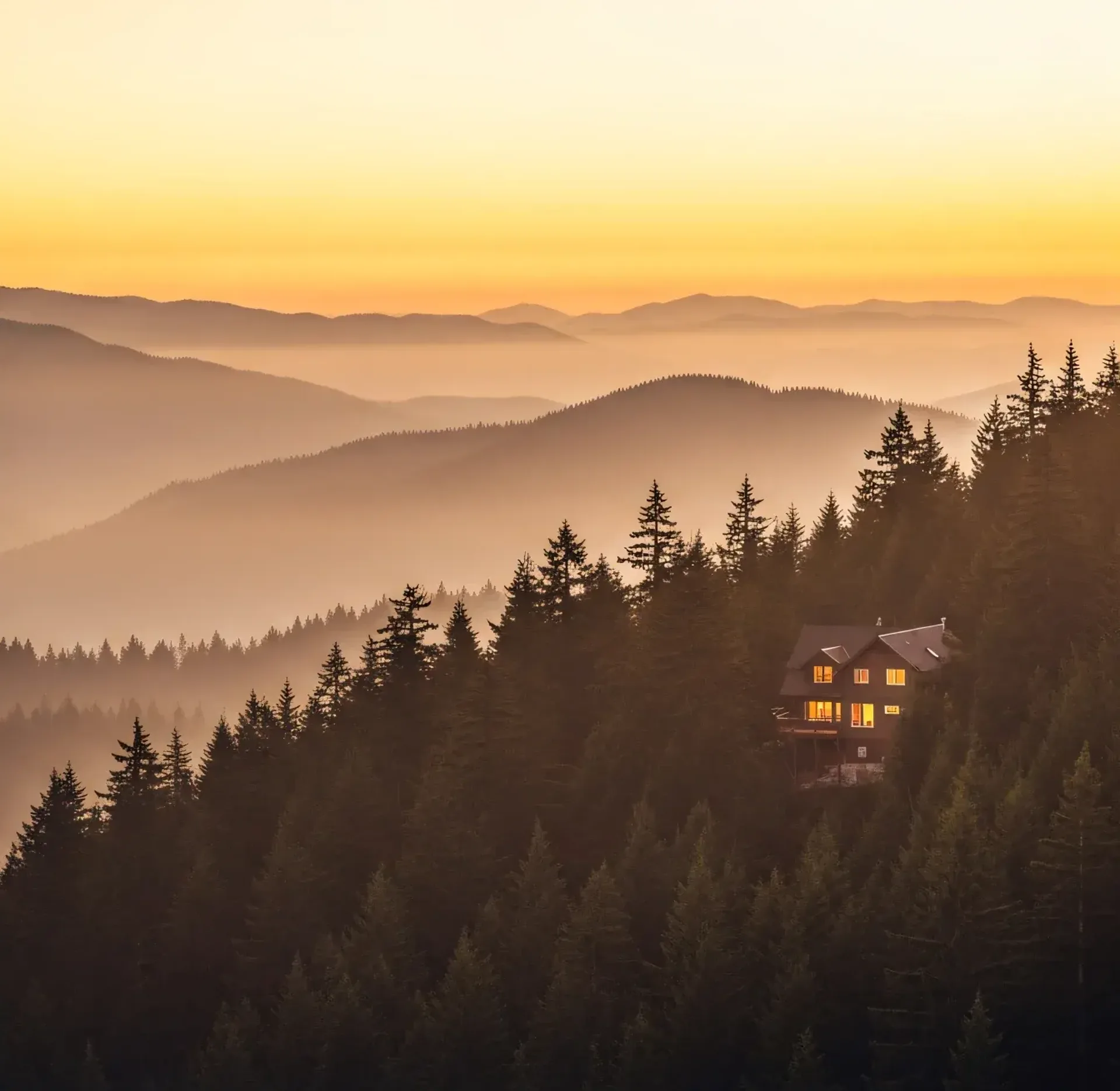 A single lit cabin on a forested ridge at dusk, layered mountain ranges fading into haze behind it, the kind of one-property rental this list is written for.